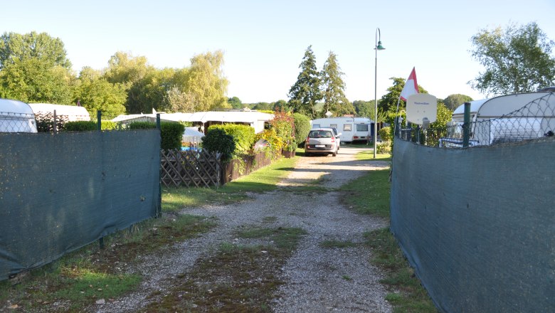 A campsite with caravans and a car on a gravel road, surrounded by trees and fences.