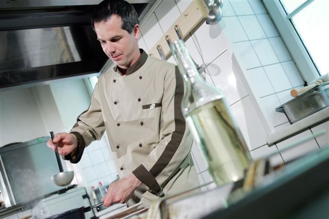 A cook in a professional kitchen scoops from a pot with a ladle.