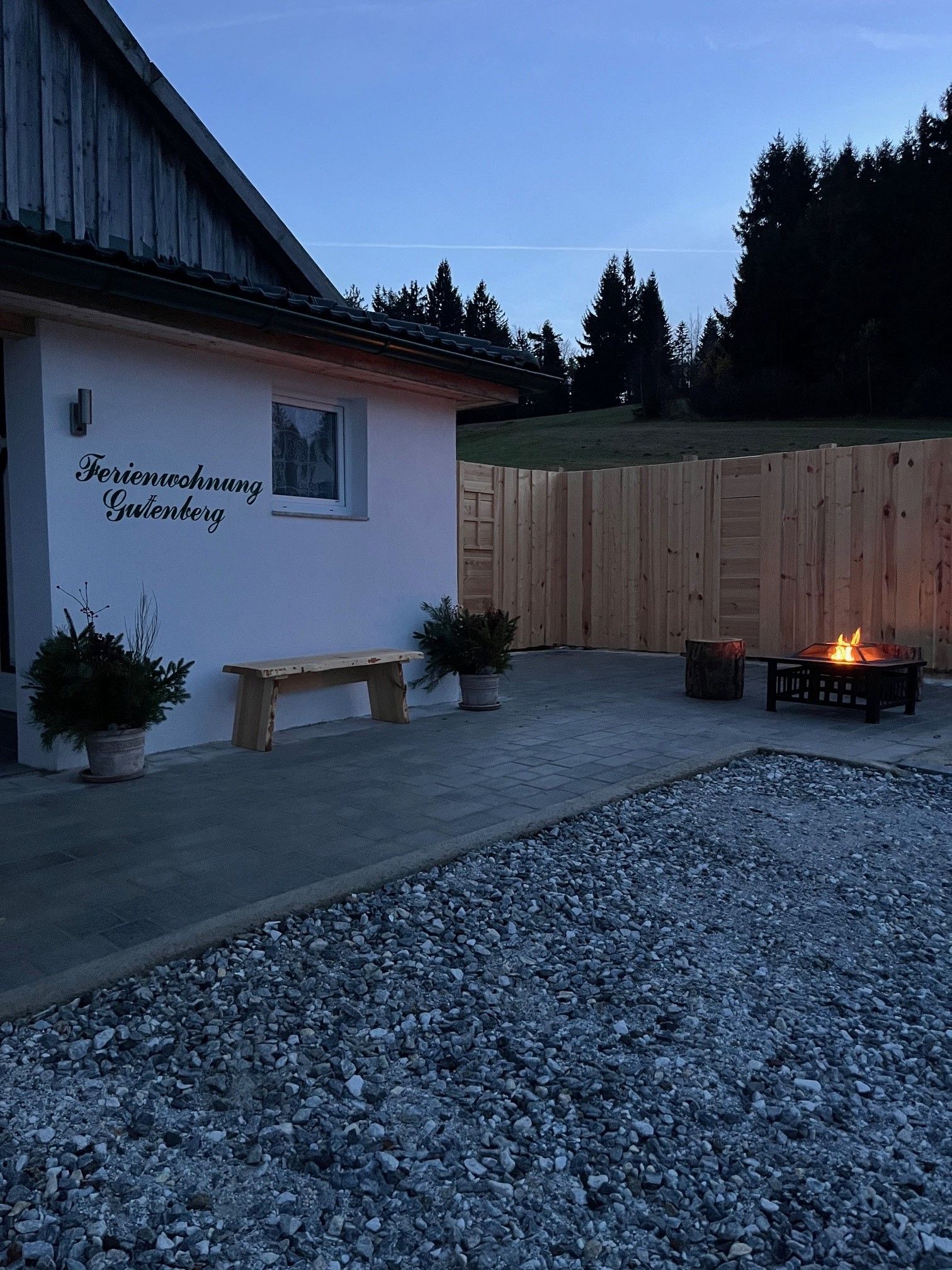 Terrace with fireplace and wooden fence at dusk, next to a building labeled 'Ferienwohnung Gutenberg'.