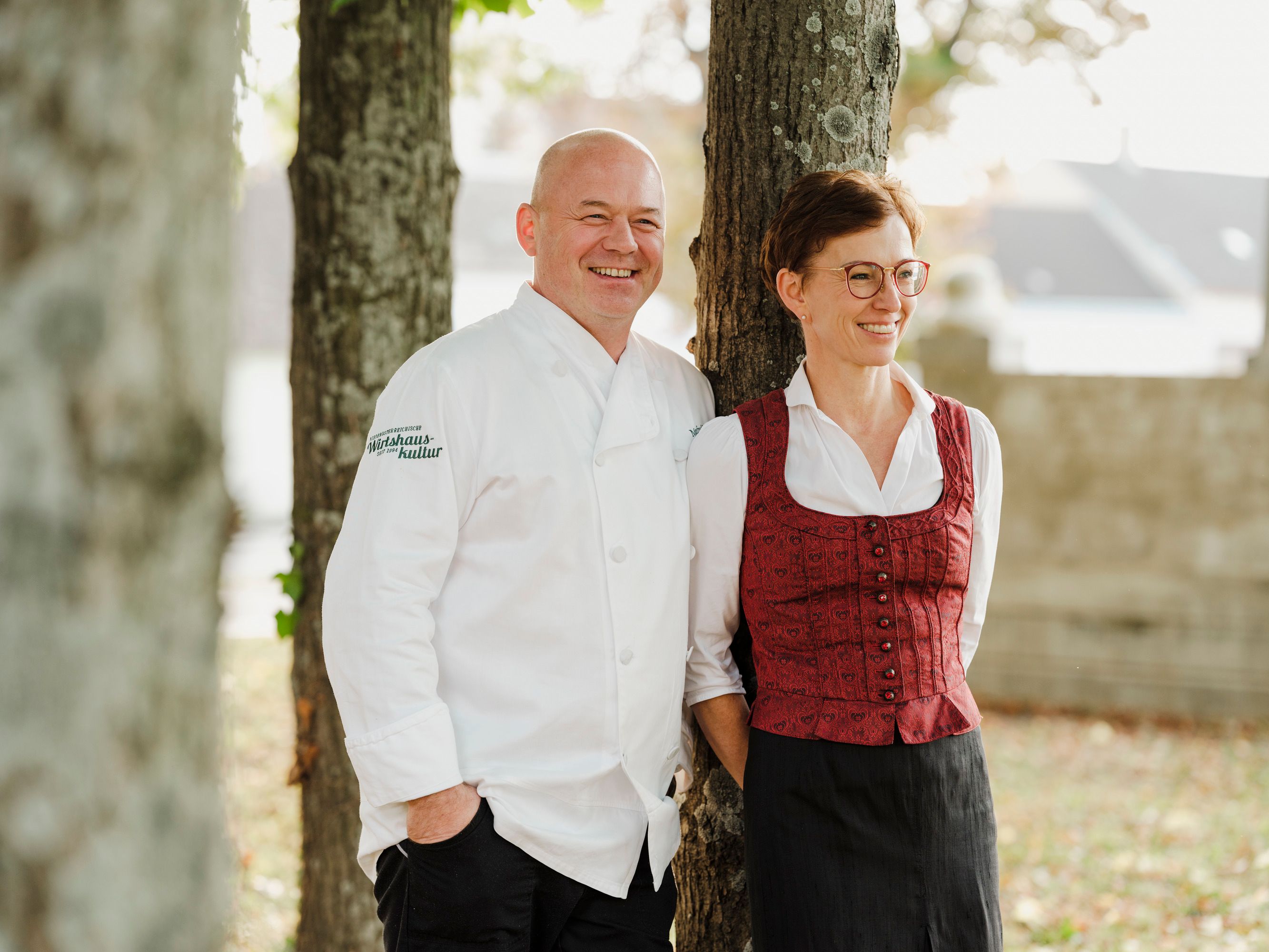 A man in a white chef's jacket and a woman in traditional dress stand smiling by a tree.