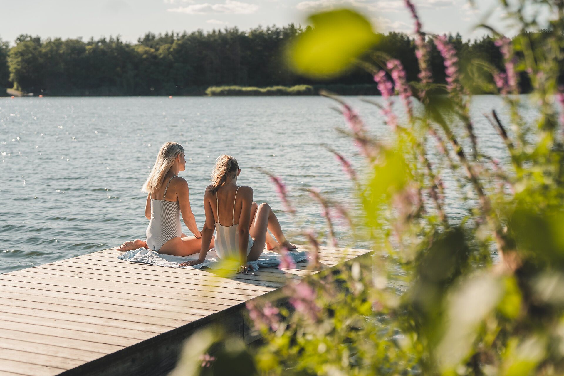 Zwei Personen in Badebekleidung sitzen auf Handtüchern auf einem Holzsteg am Viehofner See, im Hintergrund ist ein bewaldetes Ufer zu sehen.