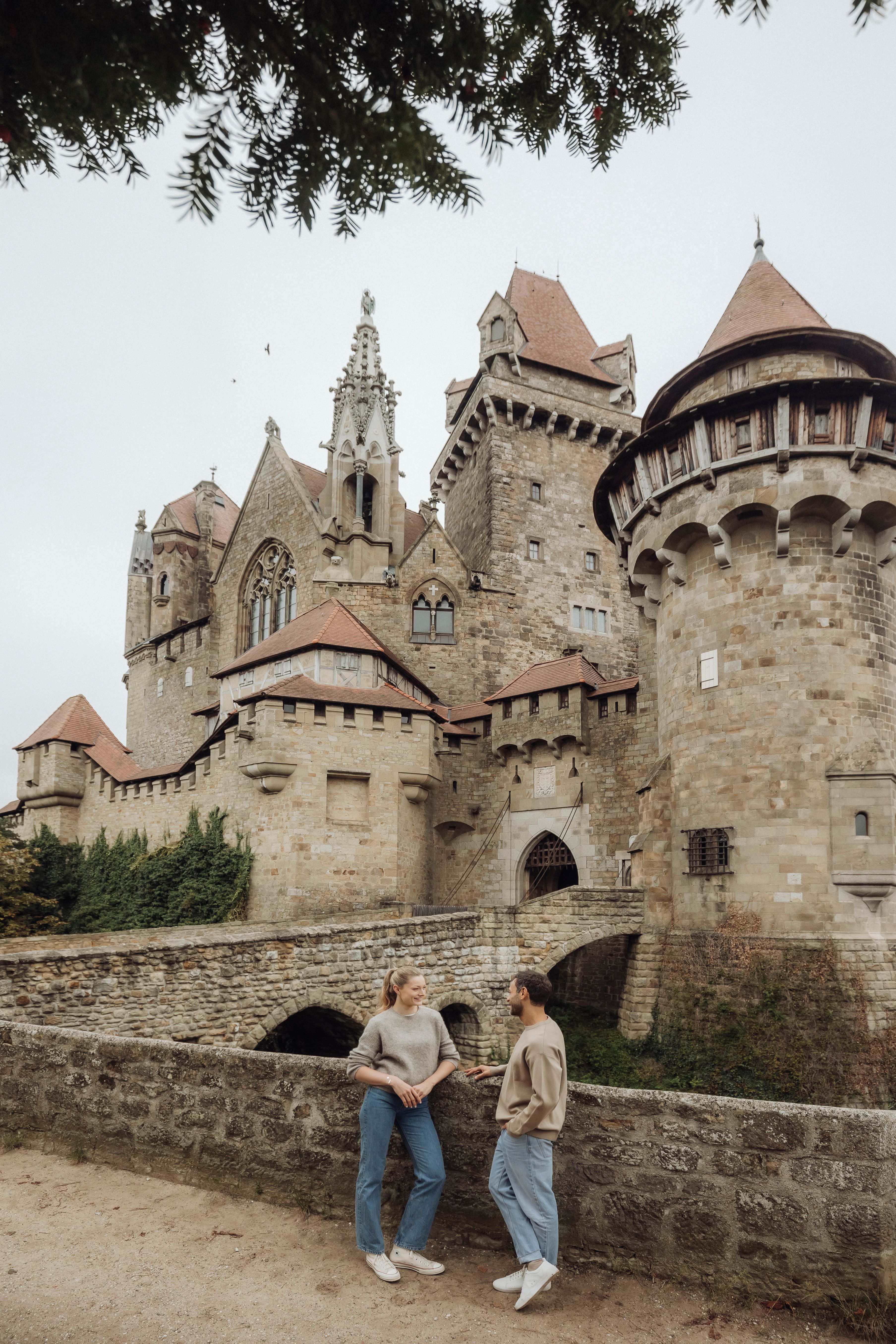 Two people are standing in front of Kreuzenstein Castle, surrounded by old stone walls and towers.