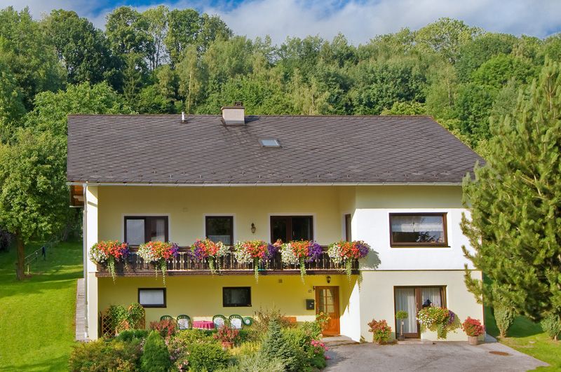 Two-storey yellow house with balcony and flowers, surrounded by trees and garden.