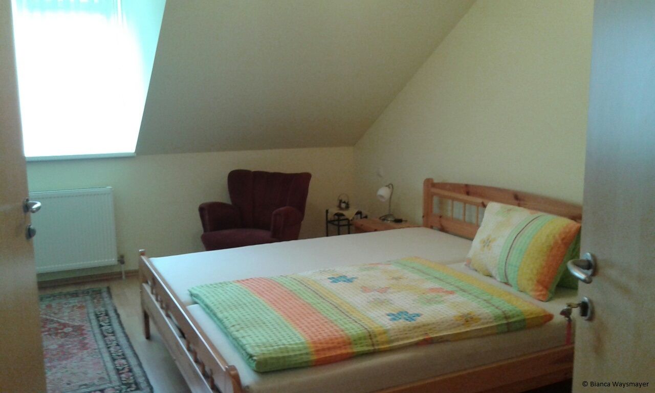A cozy bedroom with a double bed, a red armchair and a small bedside table under a sloping roof.