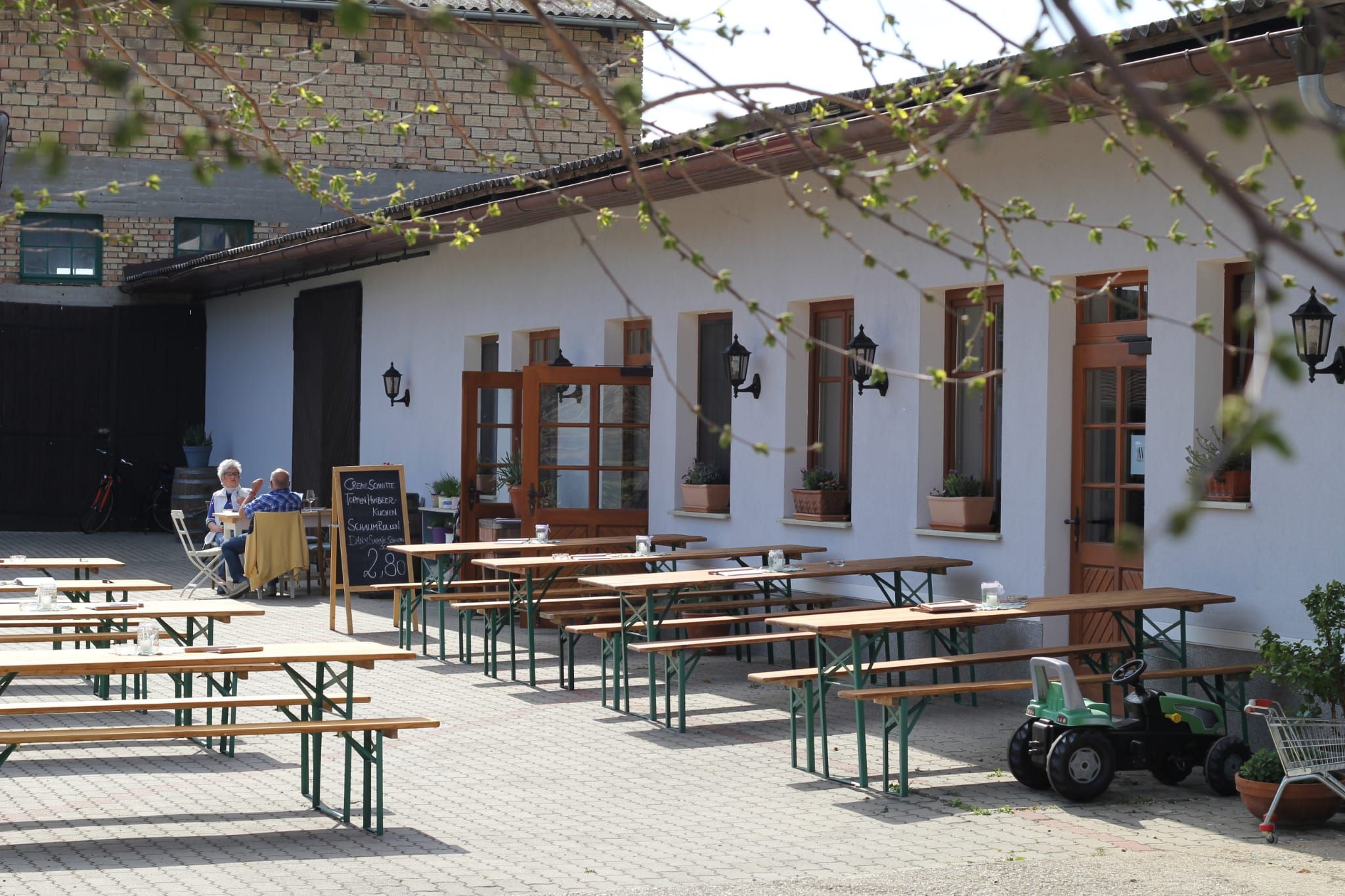 Inner courtyard of a Buschenschank (typical tavern) with empty wooden benches and a table, two people sitting at a table, a toy tractor standing next to it.