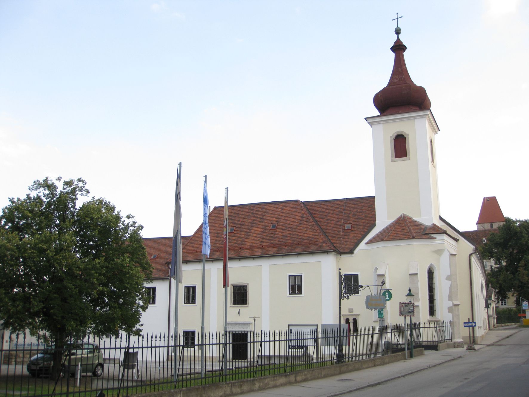 A building with a church tower and red roof tiles, surrounded by a fence and trees.