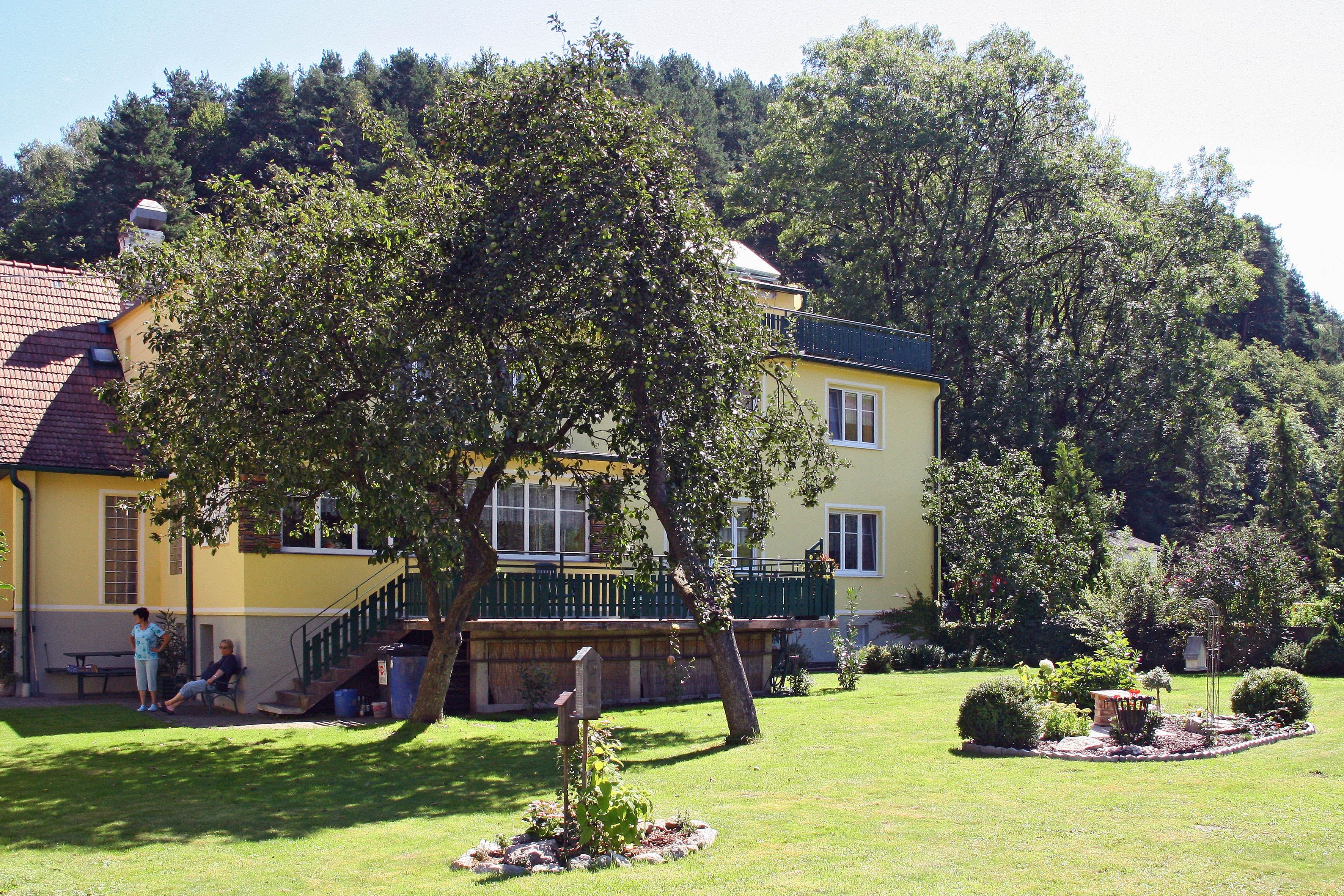 Yellow house with garden and trees, people sitting in the shade.