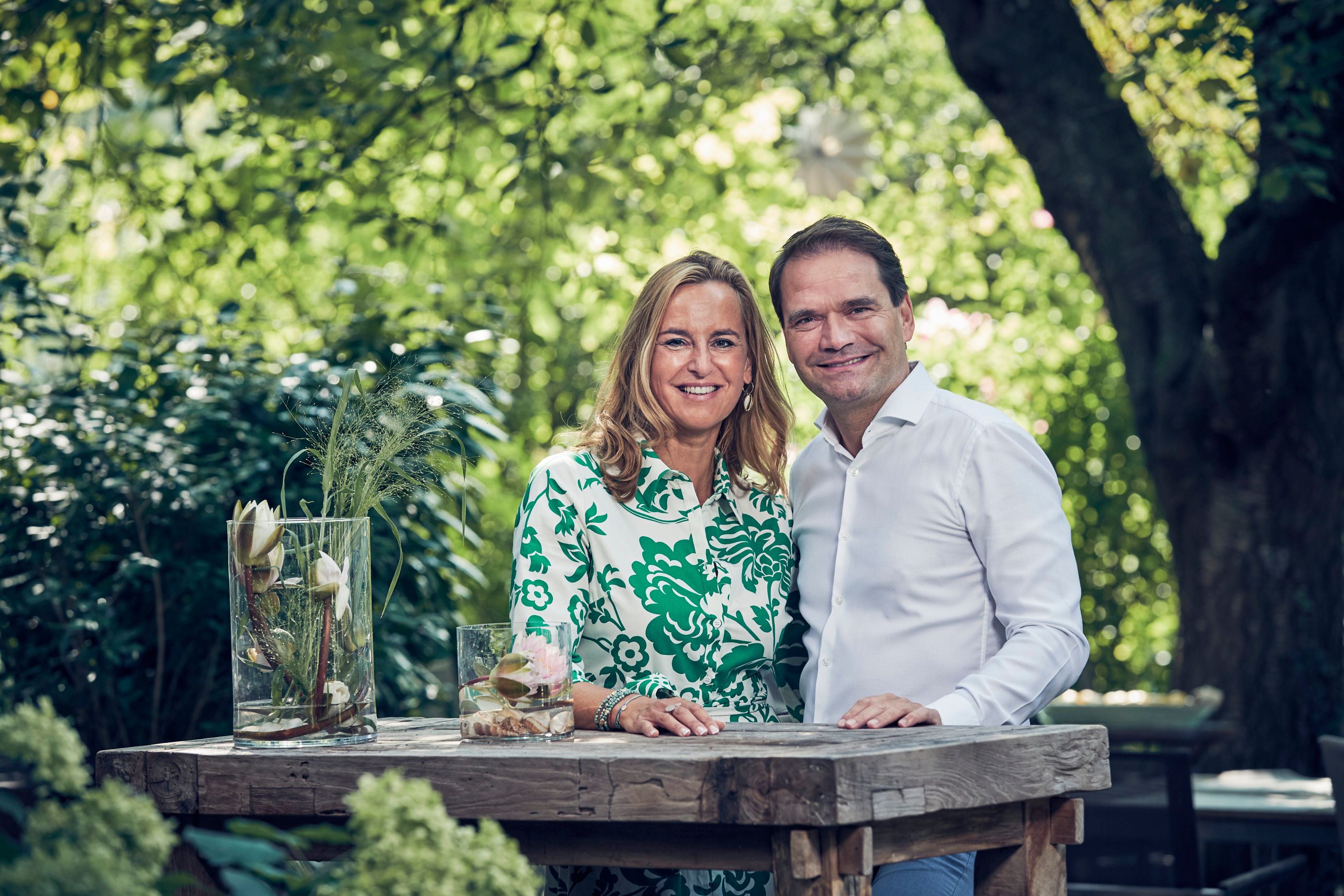 A man and a woman stand smiling at a wooden table outdoors, surrounded by green nature.