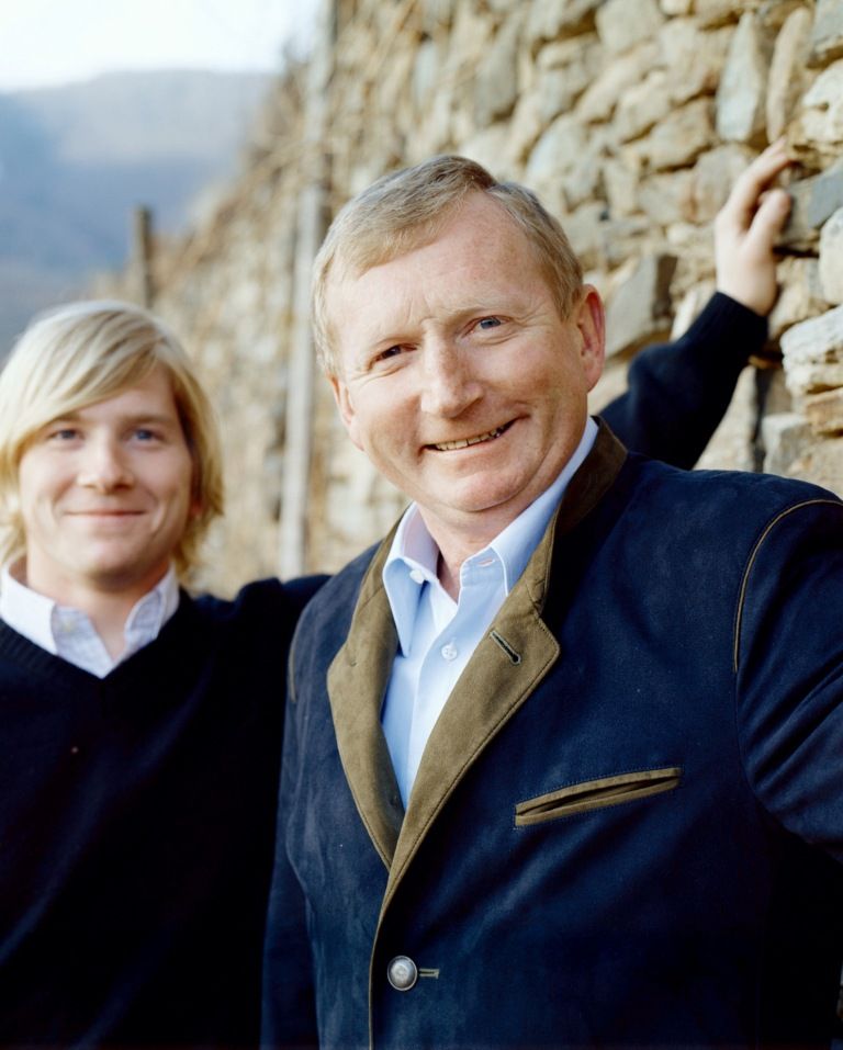 Two men stand in front of a stone wall, one smiling into the camera.