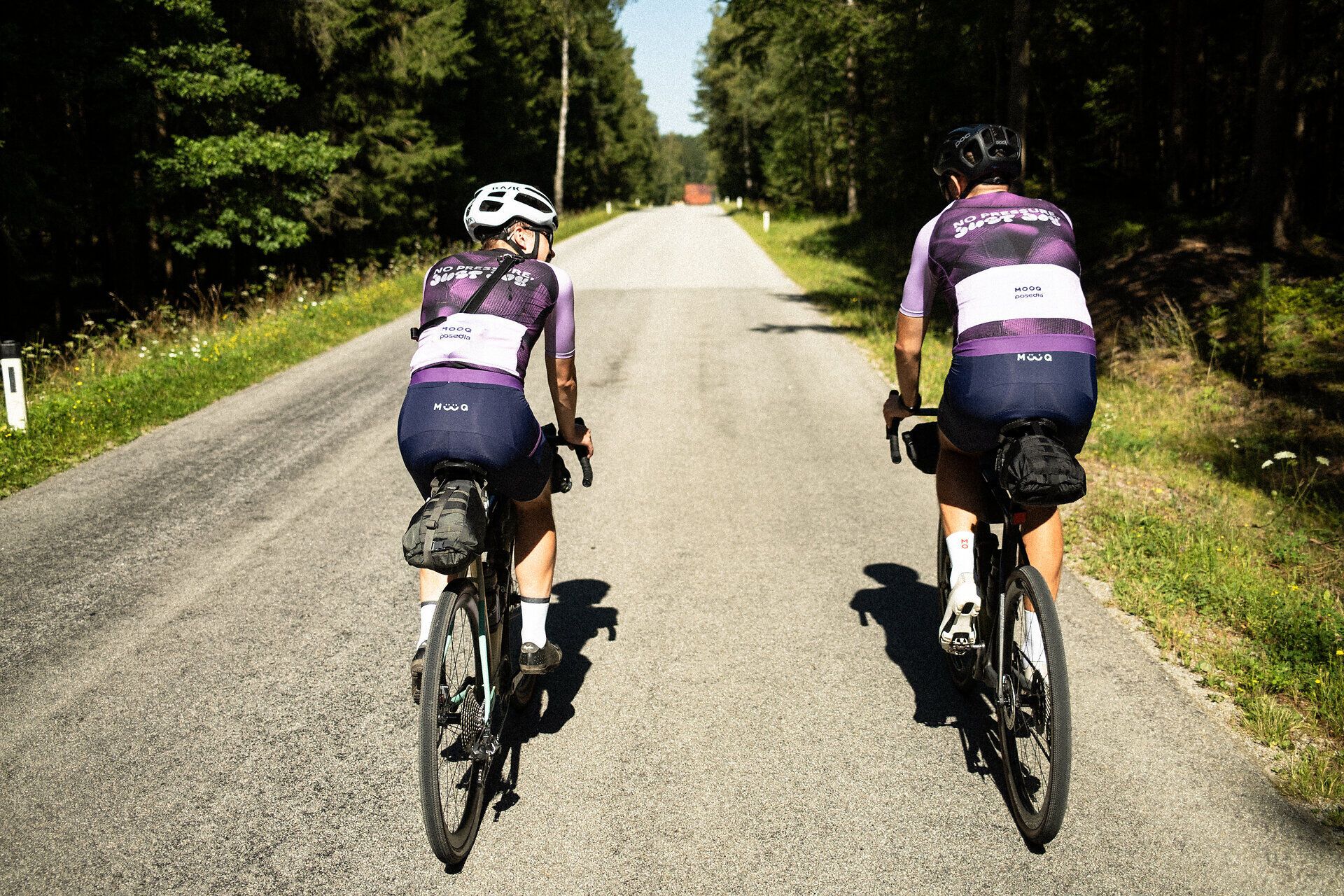 Two cyclists enjoy the fresh air and peaceful atmosphere on a picturesque country road, surrounded by lush greenery and tall trees. The rolling hills in the background invite you to explore the beauty of nature and experience the freedom of cycling.