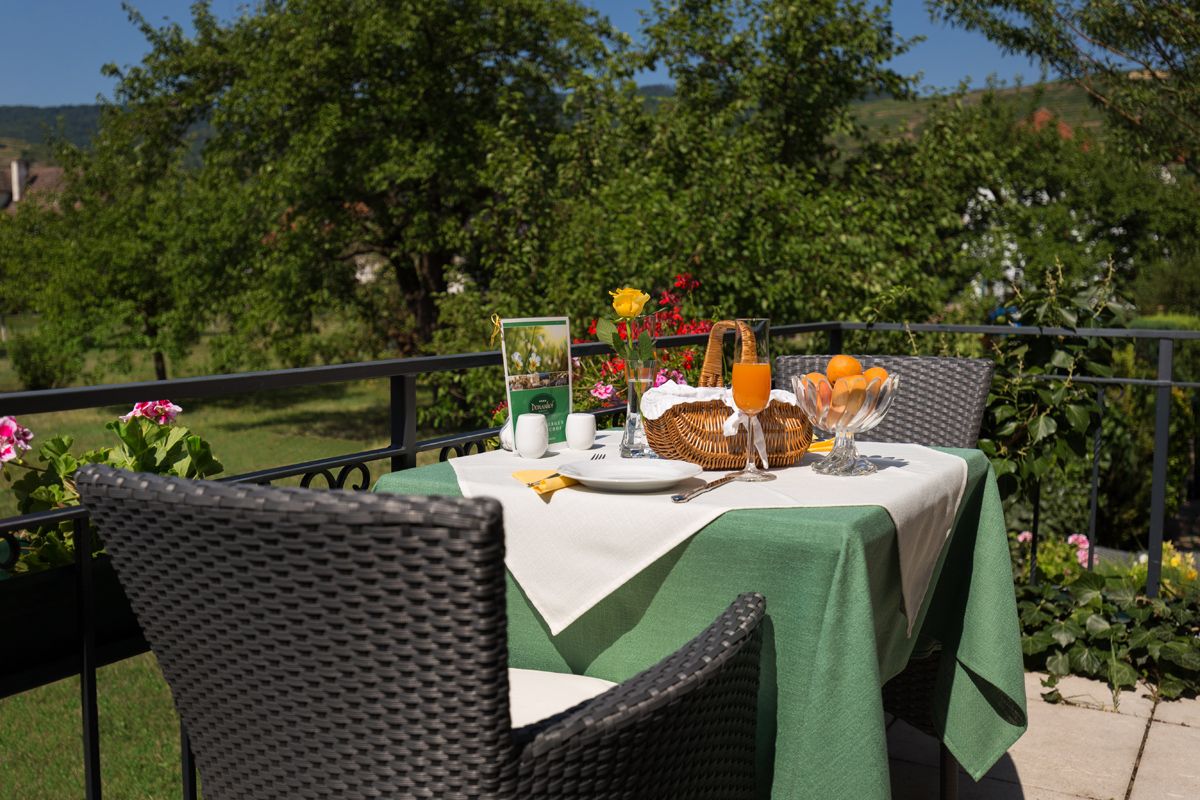 A table set on a terrace with a green tablecloth, juice, fruit and flowers, surrounded by green nature.