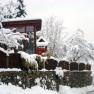 A snow-covered house with a wooden fence and stone wall, surrounded by snow-covered trees.