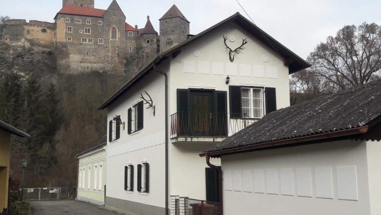 A white house with green shutters and deer antlers on the wall, with a castle on a hill in the background.