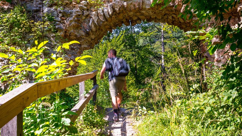 A person walks along a path under an old stone arch in the forest.