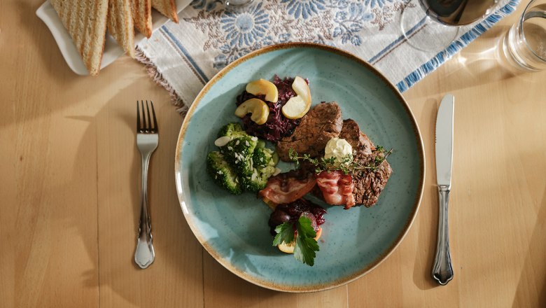 A plate of steak, broccoli, bacon and red cabbage on a table with cutlery and toast.