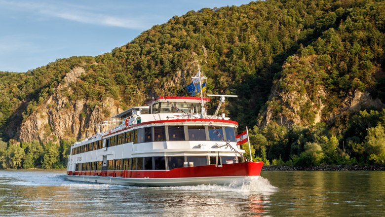 A passenger ship sails on a river against a backdrop of wooded hills.