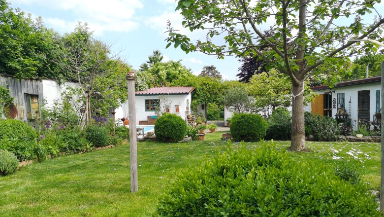 A well-tended garden with lawn, trees and shrubs, surrounded by white buildings and a blue sky in the background.