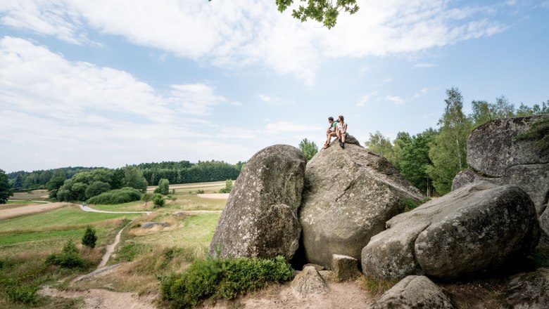 Two people are sitting on a large rock in a rural landscape with meadows and trees.