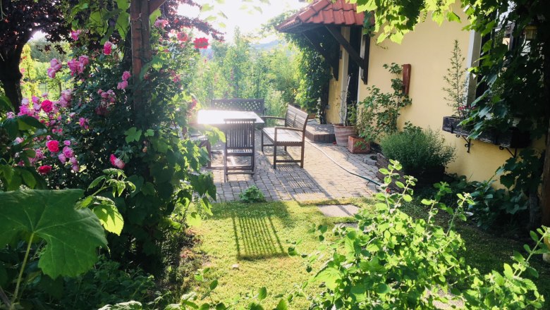 An idyllic garden with blooming roses, a table and chairs on a terrace next to a yellow house.