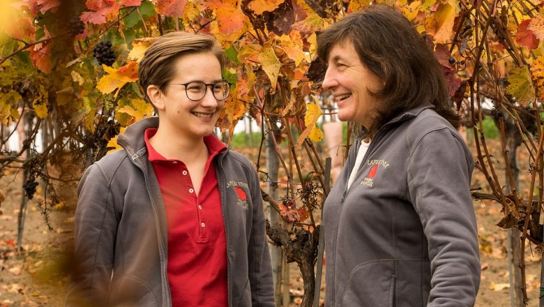 Two women stand smiling in an autumnal vineyard with colorful leaves.