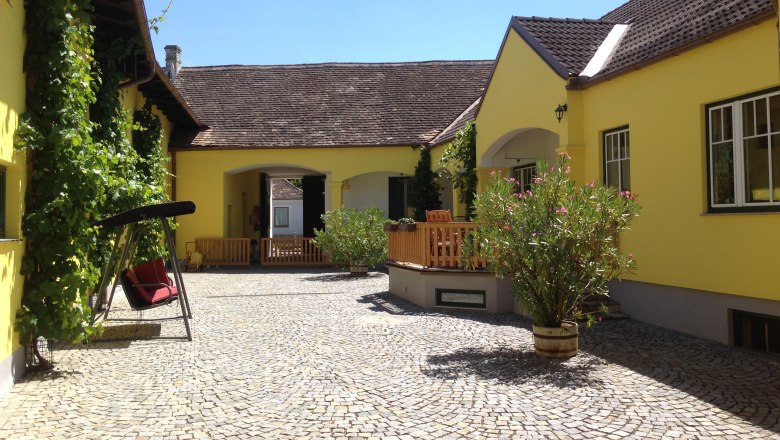 Winzerhof & Guesthouse Greil, © M. Greil Inner courtyard of a yellow vineyard with cobblestones, swing and plants.