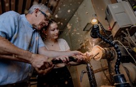 Two people work together on a lathe to machine a piece of wood. Wood shavings fly in the air.