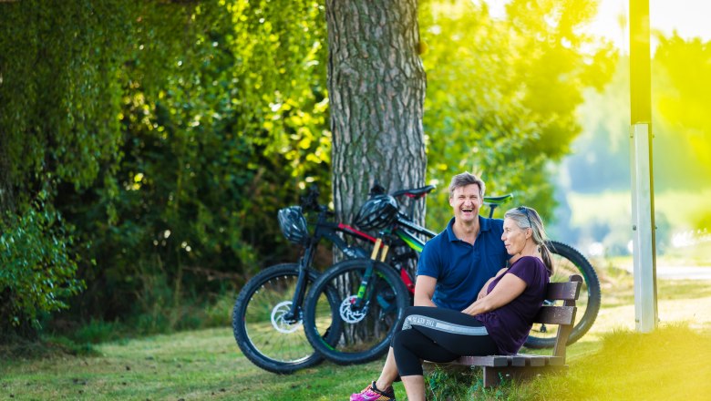 A man and a woman are sitting on a bench in the countryside, two e-bikes are parked next to them.