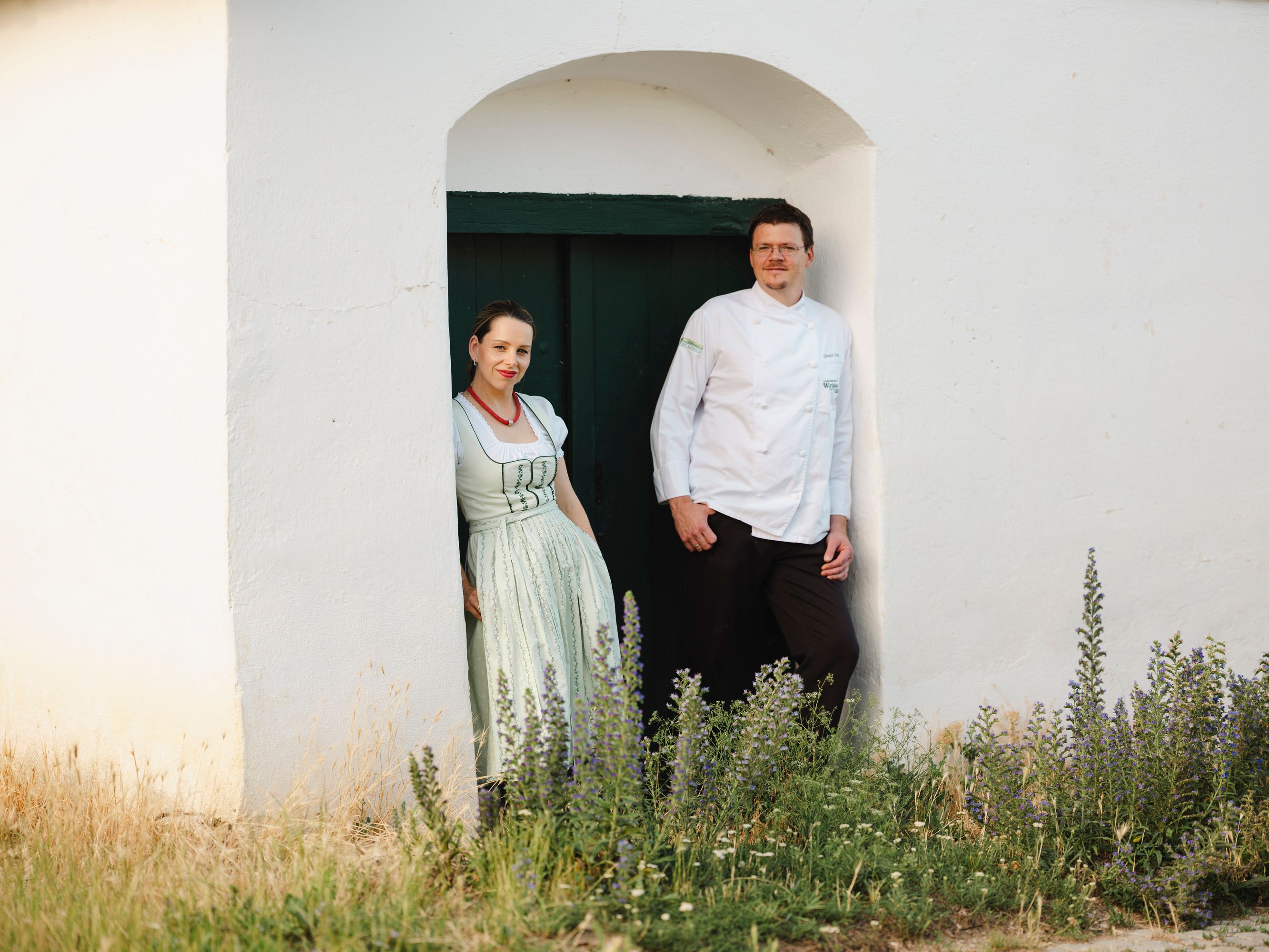 Two people are standing in a doorway in front of a white building.