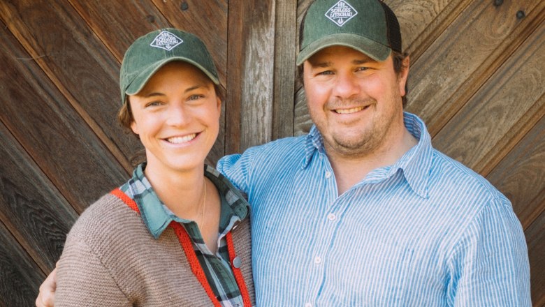 Two people are smiling at the camera, wearing green caps with a logo and standing in front of a wooden wall.