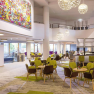 Modern hotel lobby with carpeted floor, round tables, green chairs, large lamps and a mural.