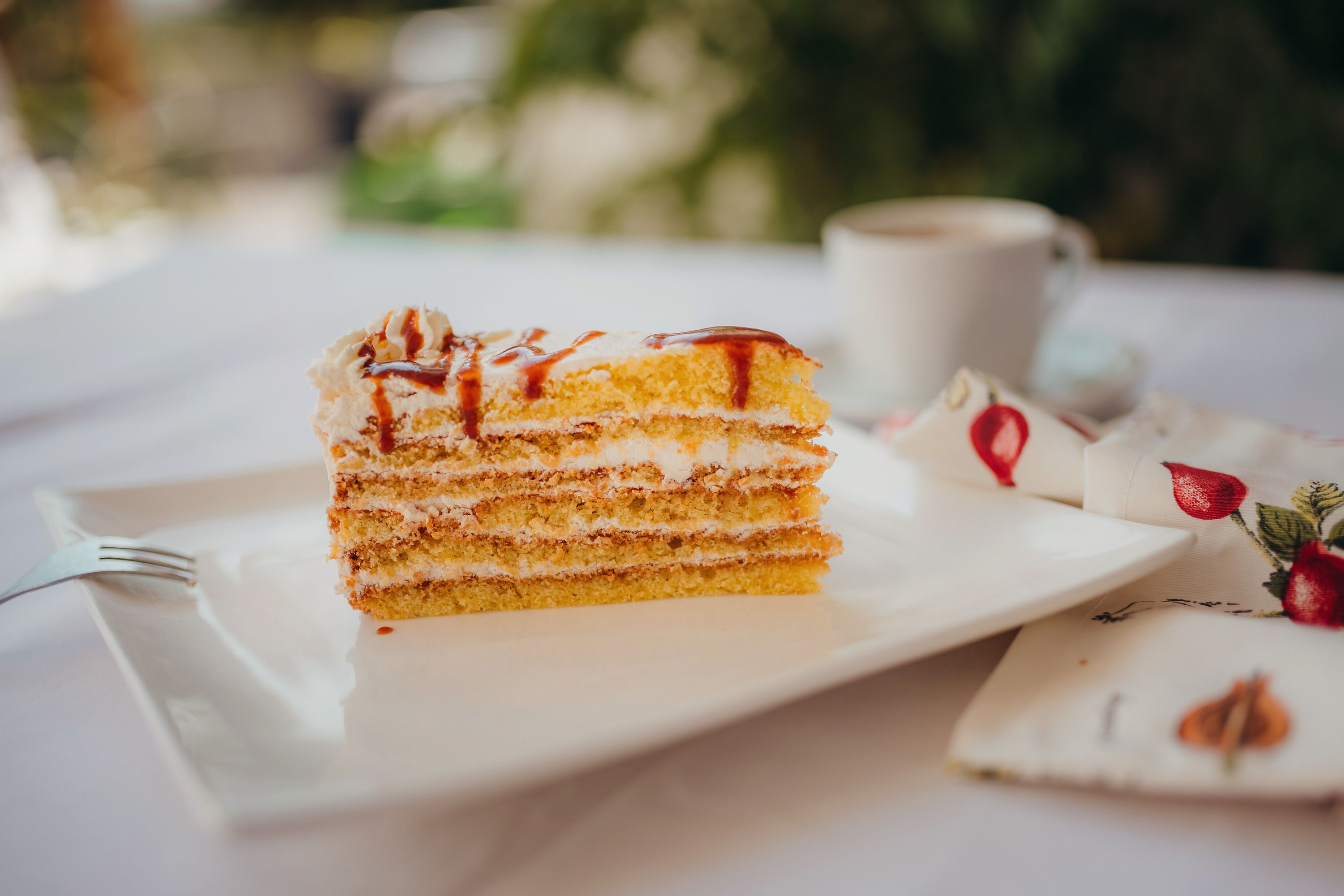 A slice of layer cake with cream and caramel on a white plate, next to it a cup of coffee and a napkin with a fruit pattern.