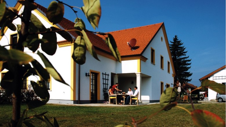Apartments Weixelbaum, © Ferienwohnungen Weixelbaum A white house with a red roof, surrounded by a green lawn and trees. People are sitting at a table outside.