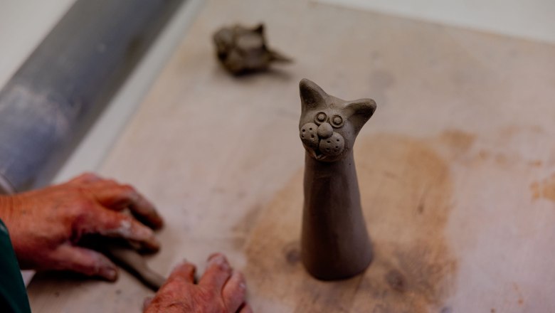 A handmade clay figure of a cat on a table, with hands shaping clay next to it.
