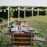 An outdoor table with wine and snacks, surrounded by fairy lights and a green landscape.
