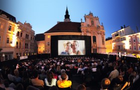 Open-air movie night in a square with a large audience in front of a big screen.