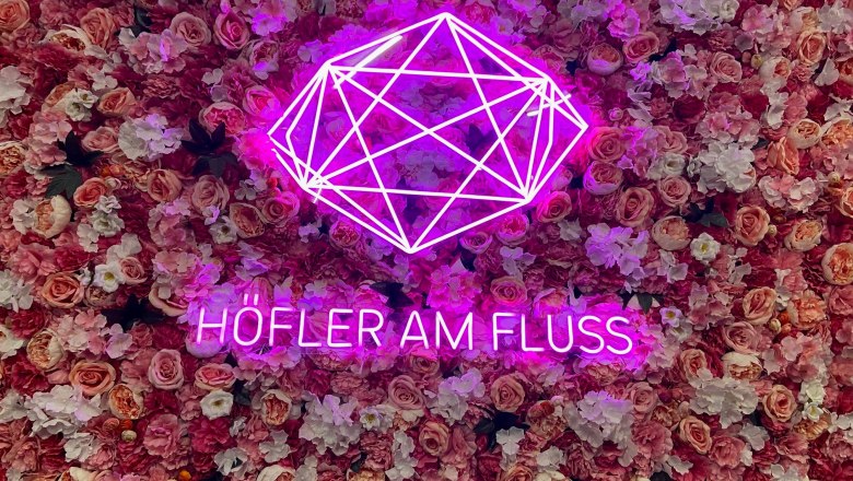 Pink neon lettering 'H&ouml;fler am Fluss' in front of a wall of pink flowers.