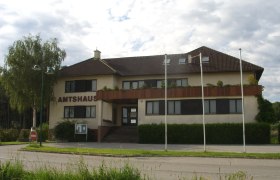 The Natschbach-Loipersbach office building with brown roof and balcony, surrounded by trees and flagpoles.