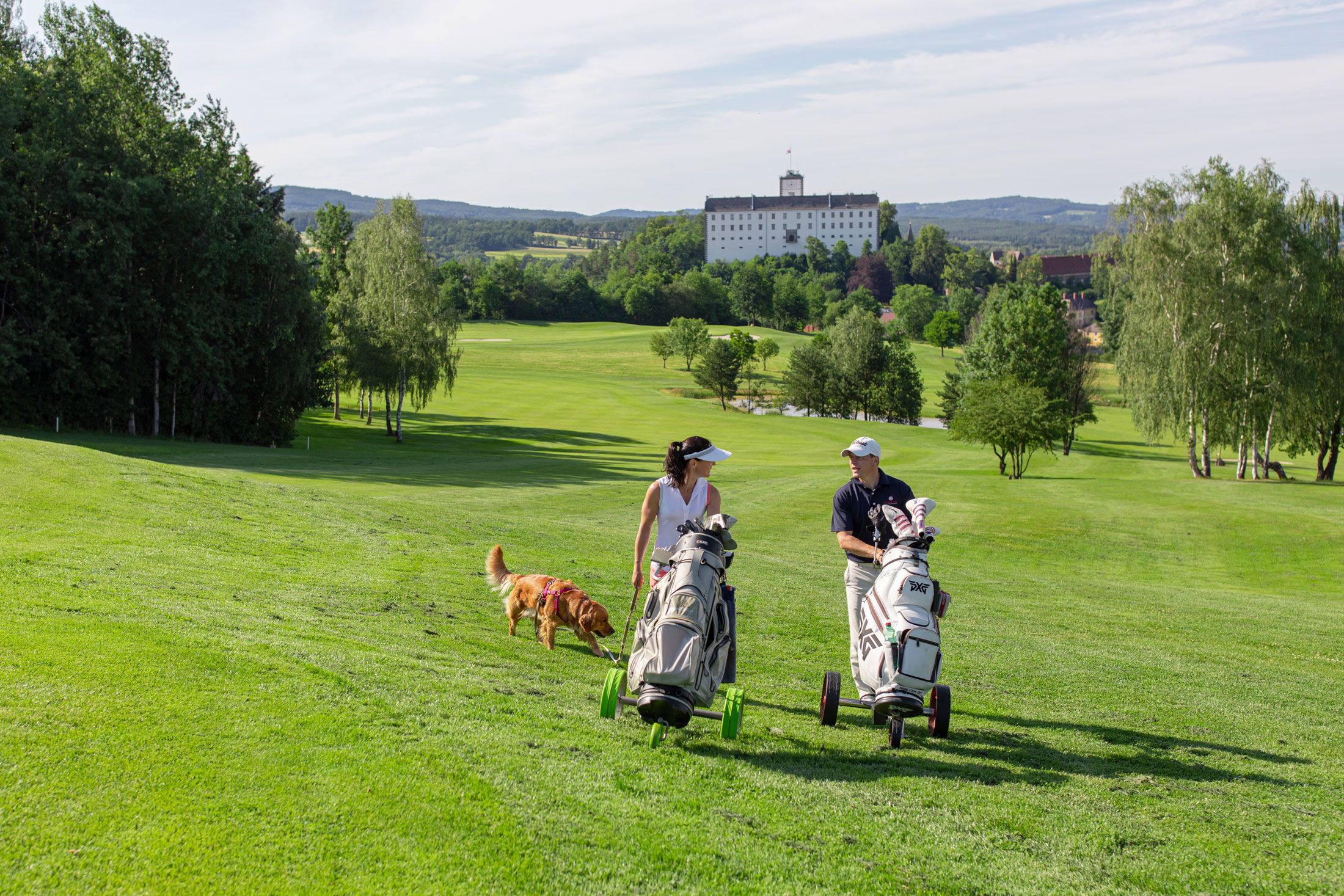 Two golfers with dog on a green golf course, castle in the background.