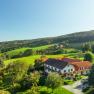 Rural landscape with a large house surrounded by green fields and trees under a blue sky.