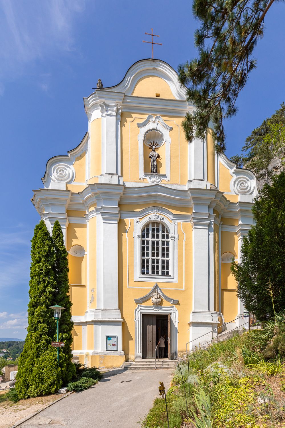 Yellow baroque church with white columns and a statue above the entrance, surrounded by trees and blue sky.
