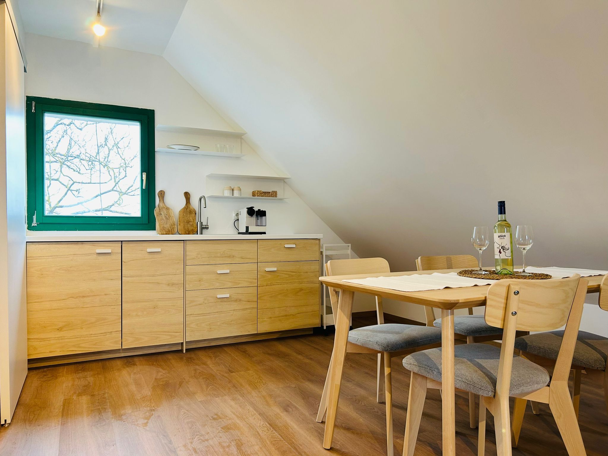 Modern kitchen with wooden cupboards, dining table and chairs, window with green frame.