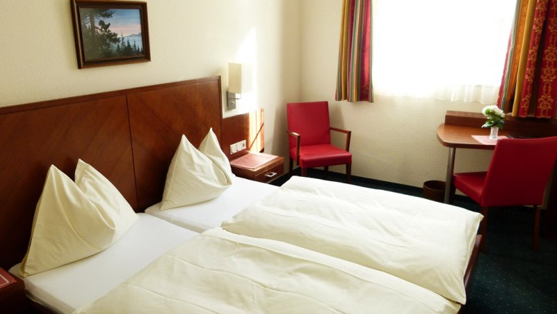 A hotel room with two single beds, red chairs and a small table by the window.