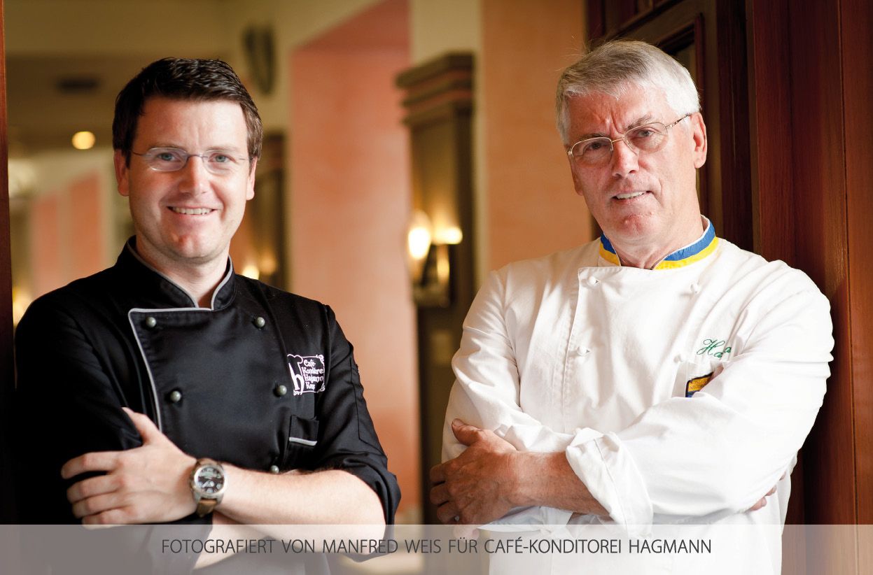 Two men in pastry chef uniforms stand next to each other and smile at the camera.