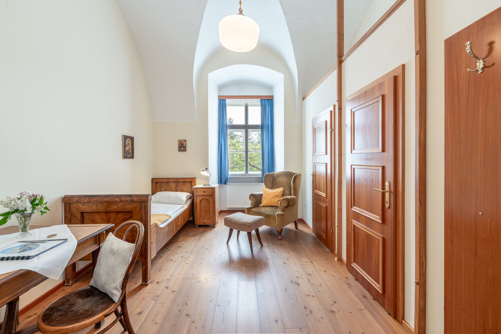 Single room in Altenburg Abbey with bed, armchair, table and window with blue curtains.