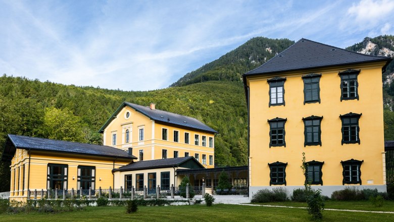 Exterior view of the yellow Thalhof in front of wooded mountains.