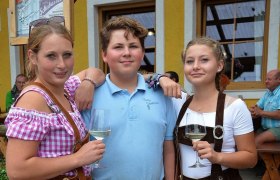 Three young people in traditional Bavarian dress with glasses of wine in front of a yellow building.