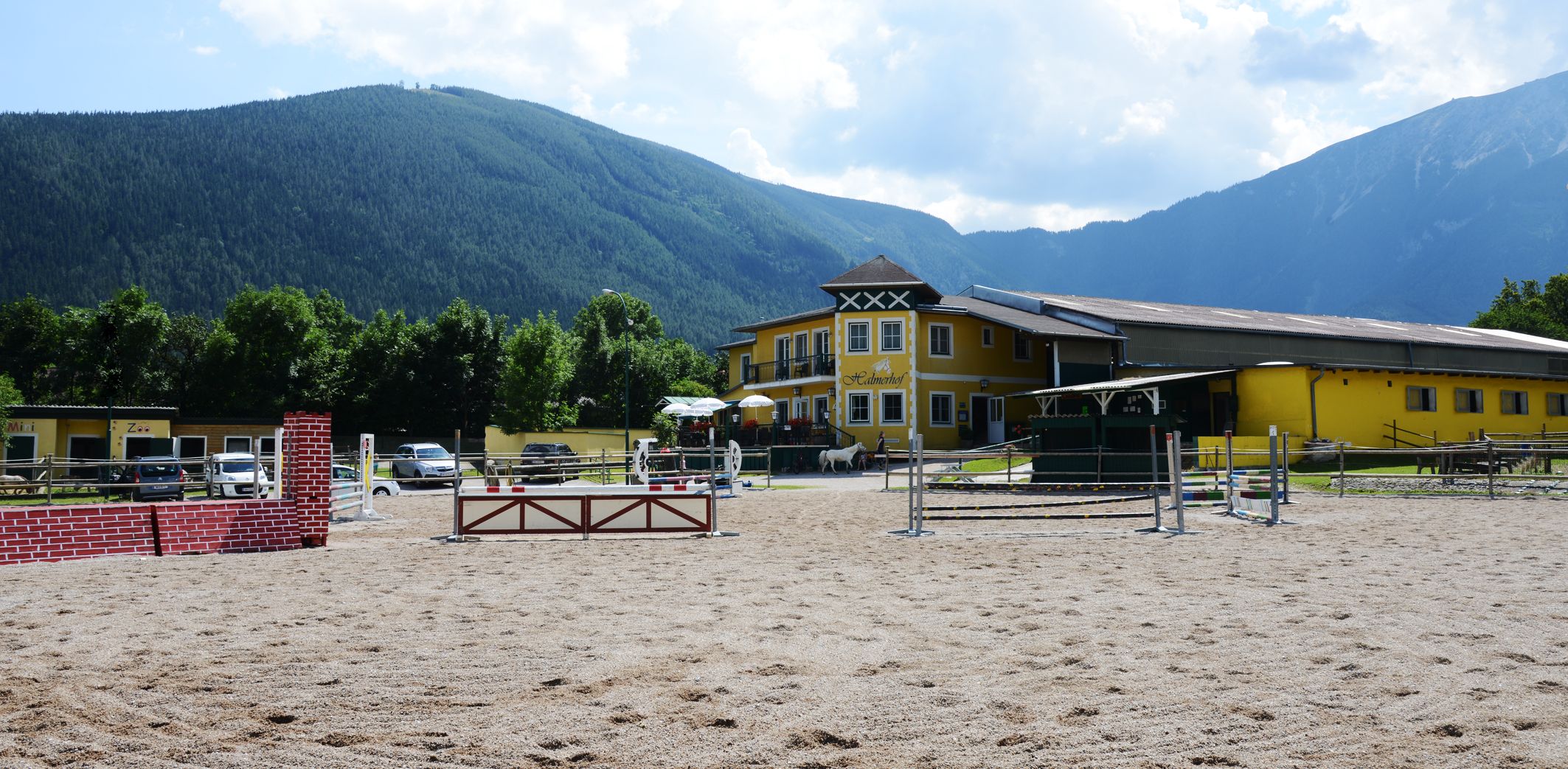 Halmerhof riding stables with jumping arena and mountains in the background.