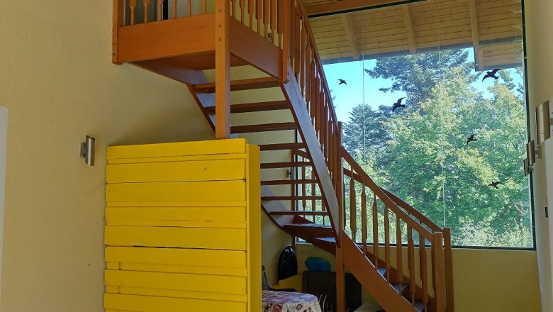 Wooden staircase in an interior with a yellow wall and large window with a view of trees.