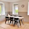 Dining area with wooden table and black chairs, light-colored walls, large windows, clock on the wall.
