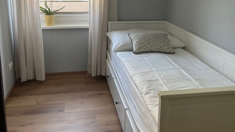 Small bedroom with single bed, white curtains and a plant on the windowsill.